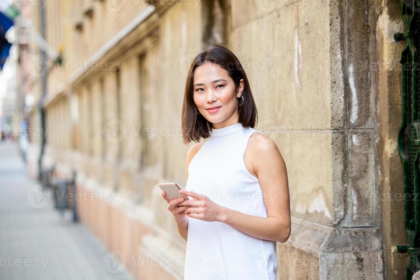 Beautiful asian woman using an application to send an sms message in her smartphone device. Happy young Asian woman with smartphone standing in the street photo