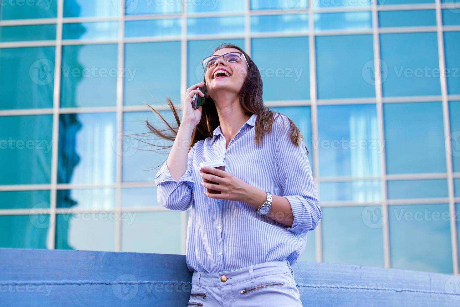 Happy smiling business woman talking on mobile smartphone and drinking coffee from disposable paper cup in the street with office buildings in the background photo