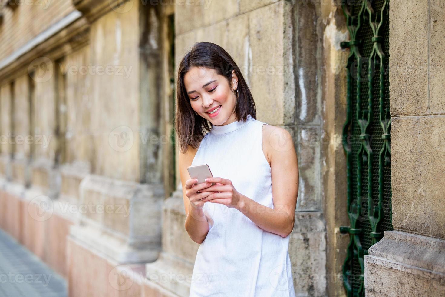Beautiful asian woman using an application to send an sms message in her smartphone device. Happy young Asian woman with smartphone standing in the street photo