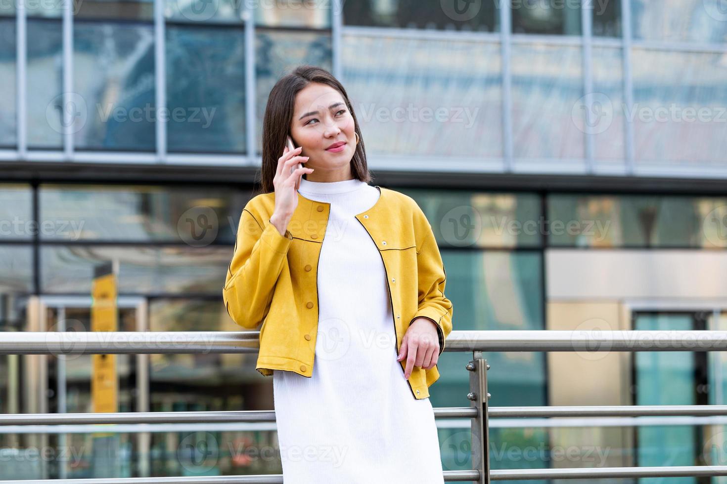 Happy smiling business woman talking on mobile smartphone in the street with office buildings in the background. Executive business Asian woman talking on mobile cellphone photo