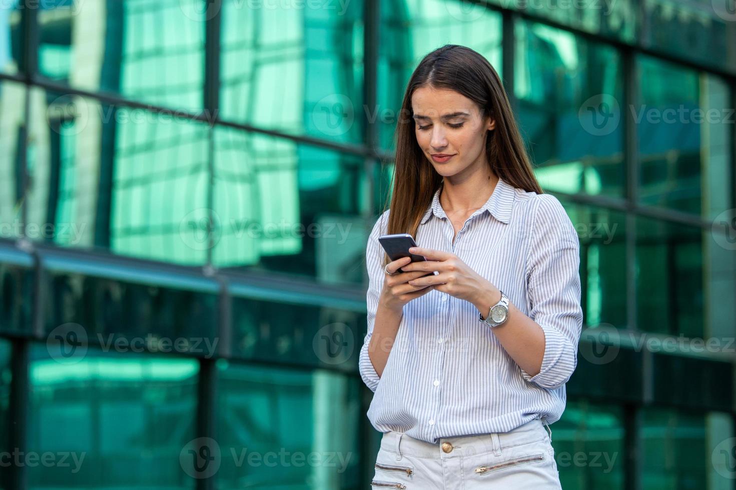 Executive business woman talking on mobile smartphone in the street with office buildings in the background. Young woman with smartphone standing against street blurred building background. photo