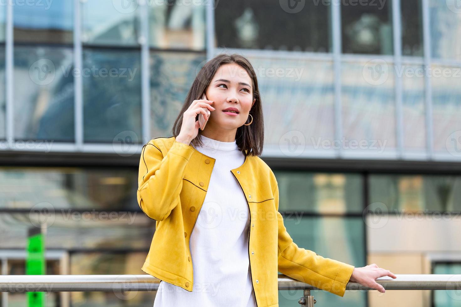 Happy smiling business woman talking on mobile smartphone in the street with office buildings in the background. Executive business Asian woman talking on mobile smartphone photo