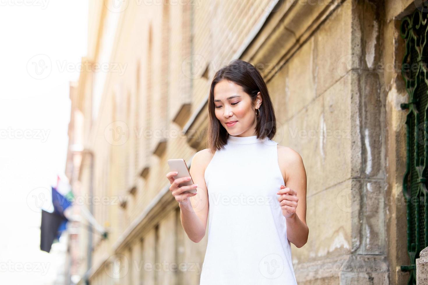 Beautiful asian woman using an application to send an sms message in her smartphone device. Happy young Asian woman with smartphone standing in the street photo