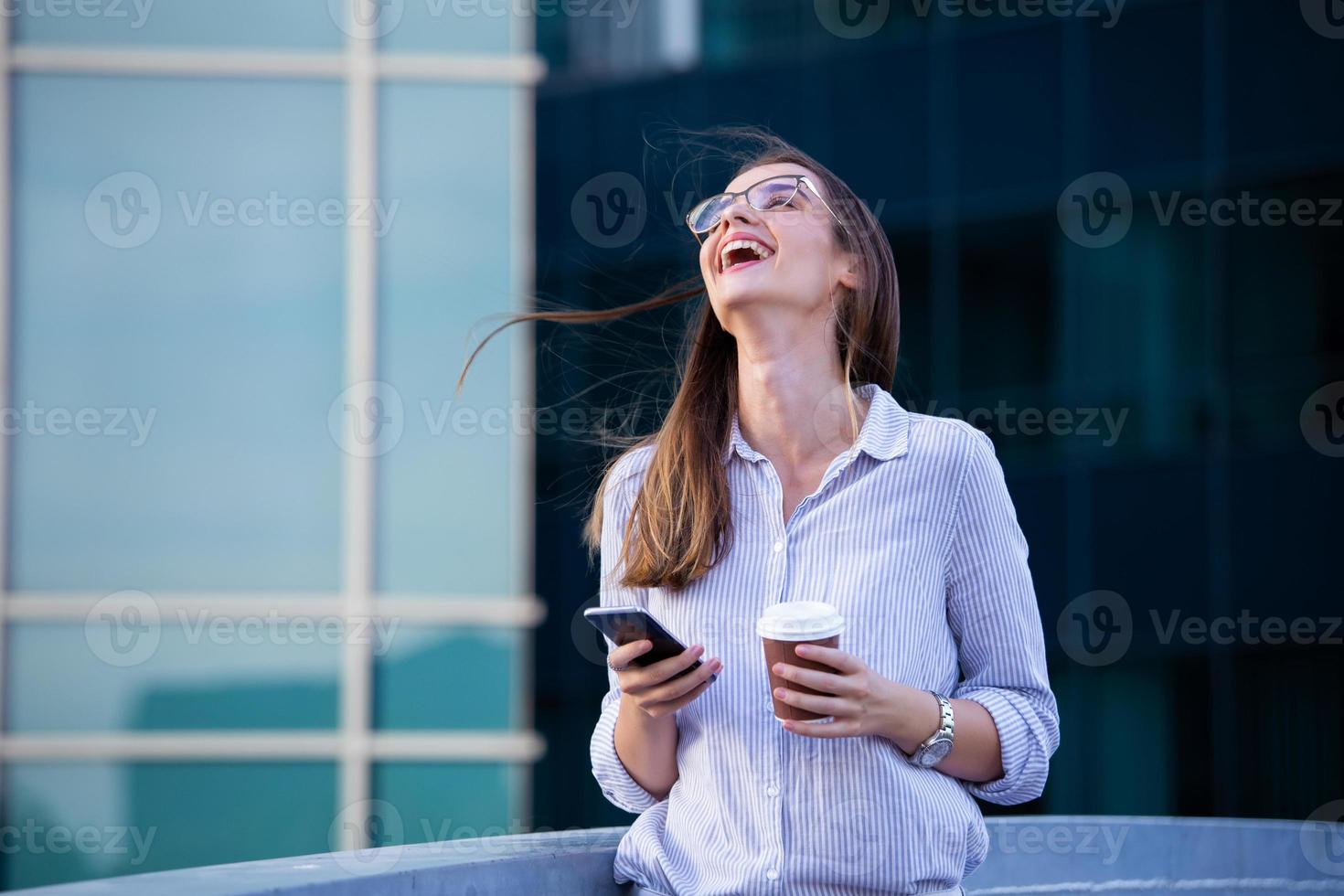 Happy smiling business woman on mobile smartphone and drinking coffee from disposable paper cup in the street with office buildings in the background photo