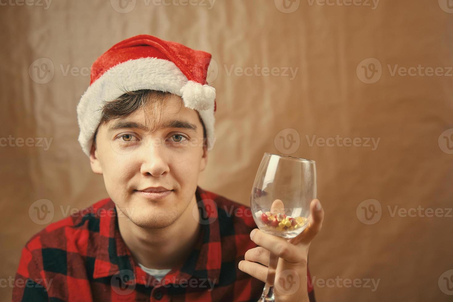 Guy in Santa Claus hat holds a glass with various pills. photo
