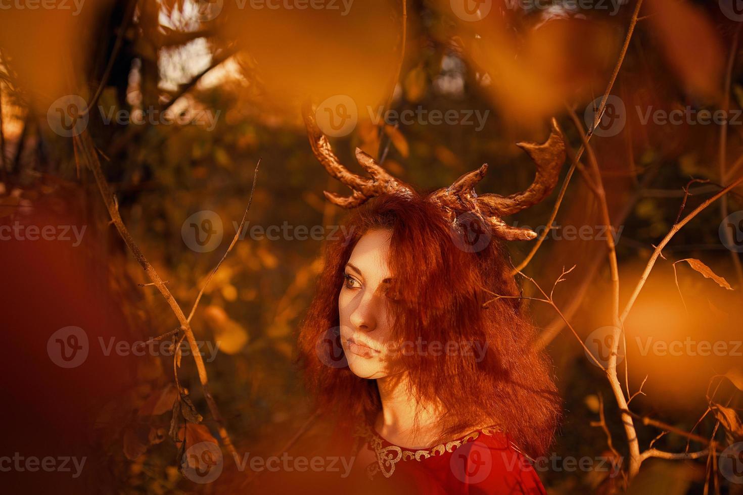 Woman in long red dress with deer horns in autumn forest. photo