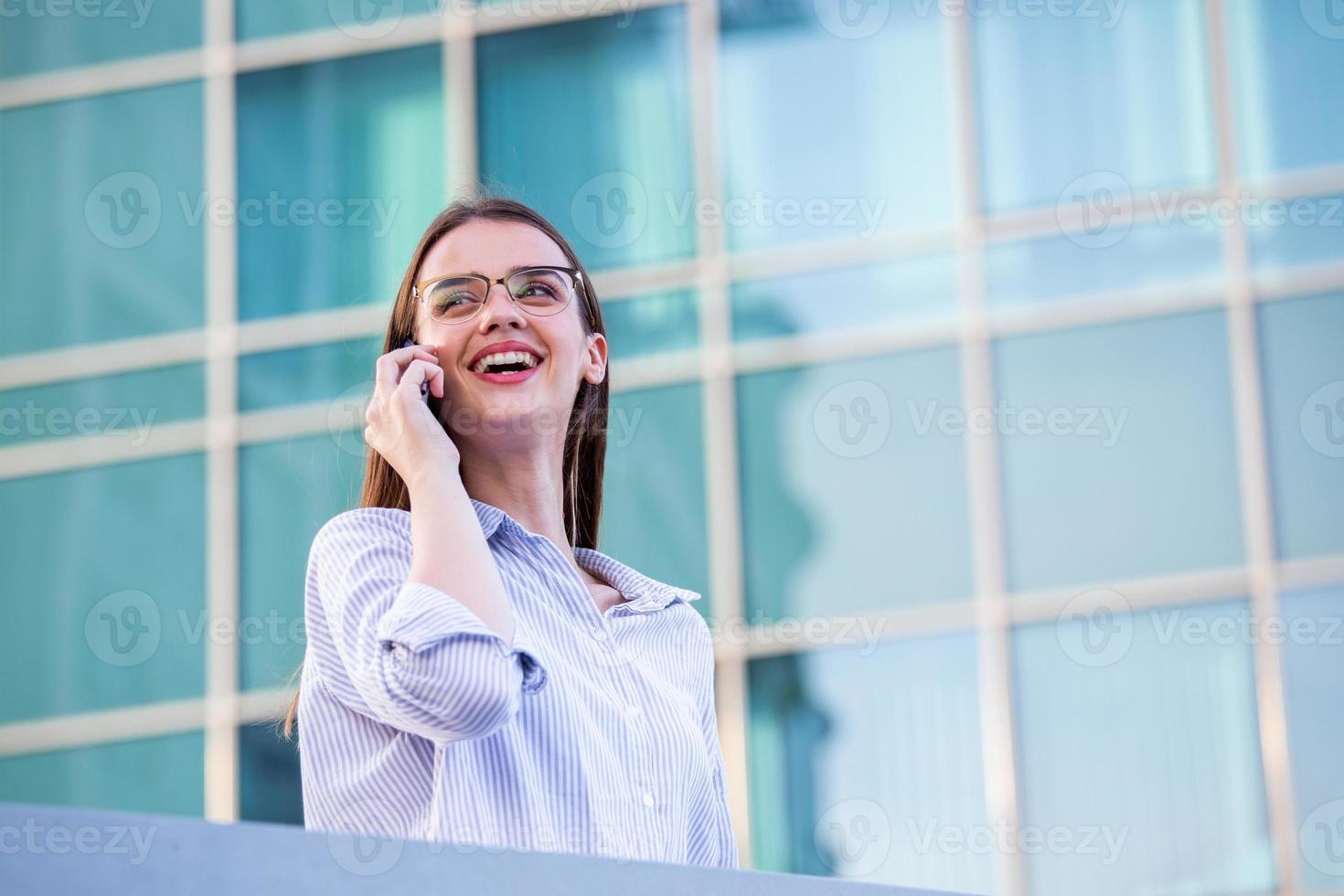 Happy smiling business woman talking on mobile smartphone in the street with office buildings in the background photo