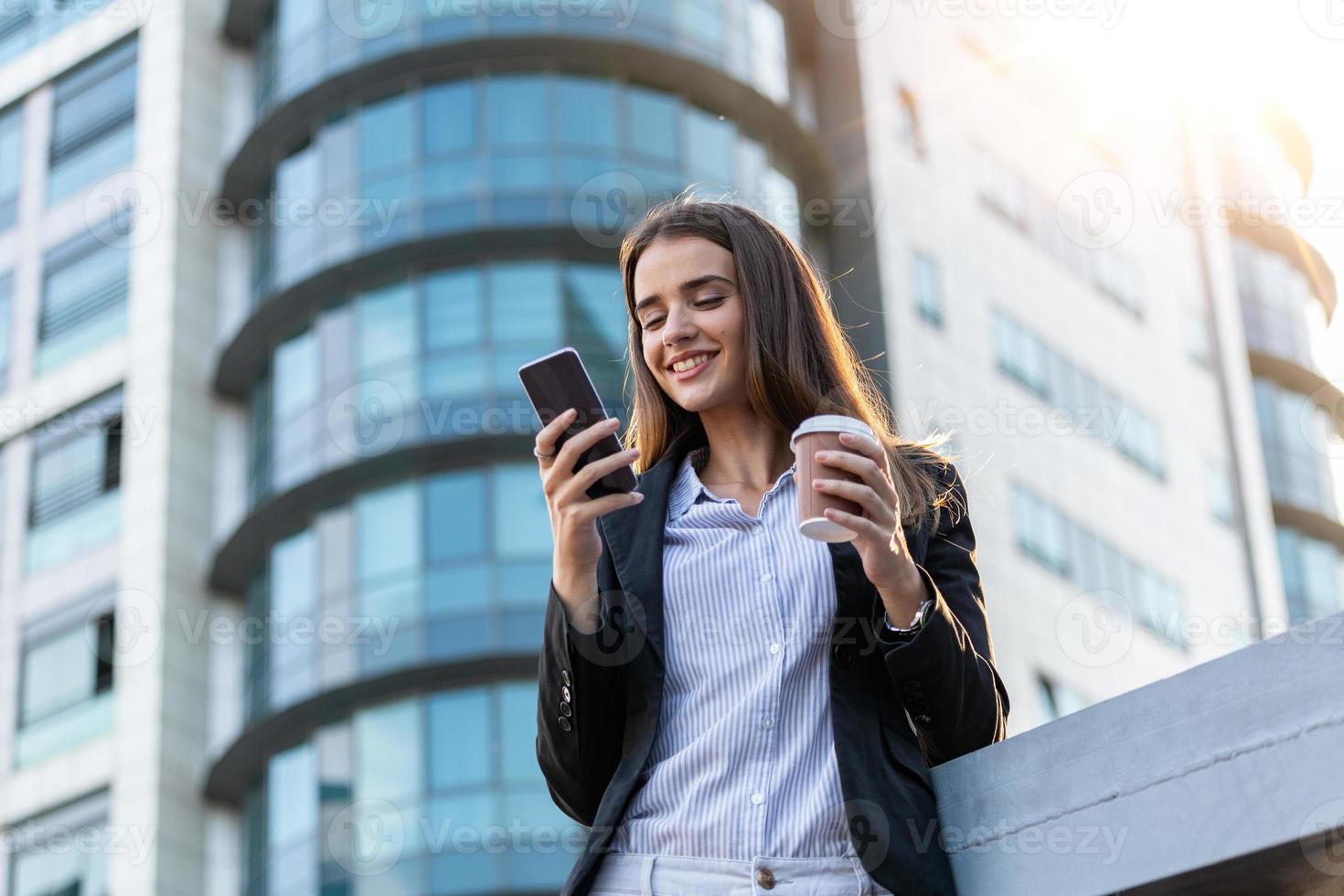 mujer de negocios con café y hablando por teléfono cerca de la oficina. mujer joven con smartphone de pie contra la calle fondo de edificio borroso. hermosa chica en suite con teléfono y taza de café foto