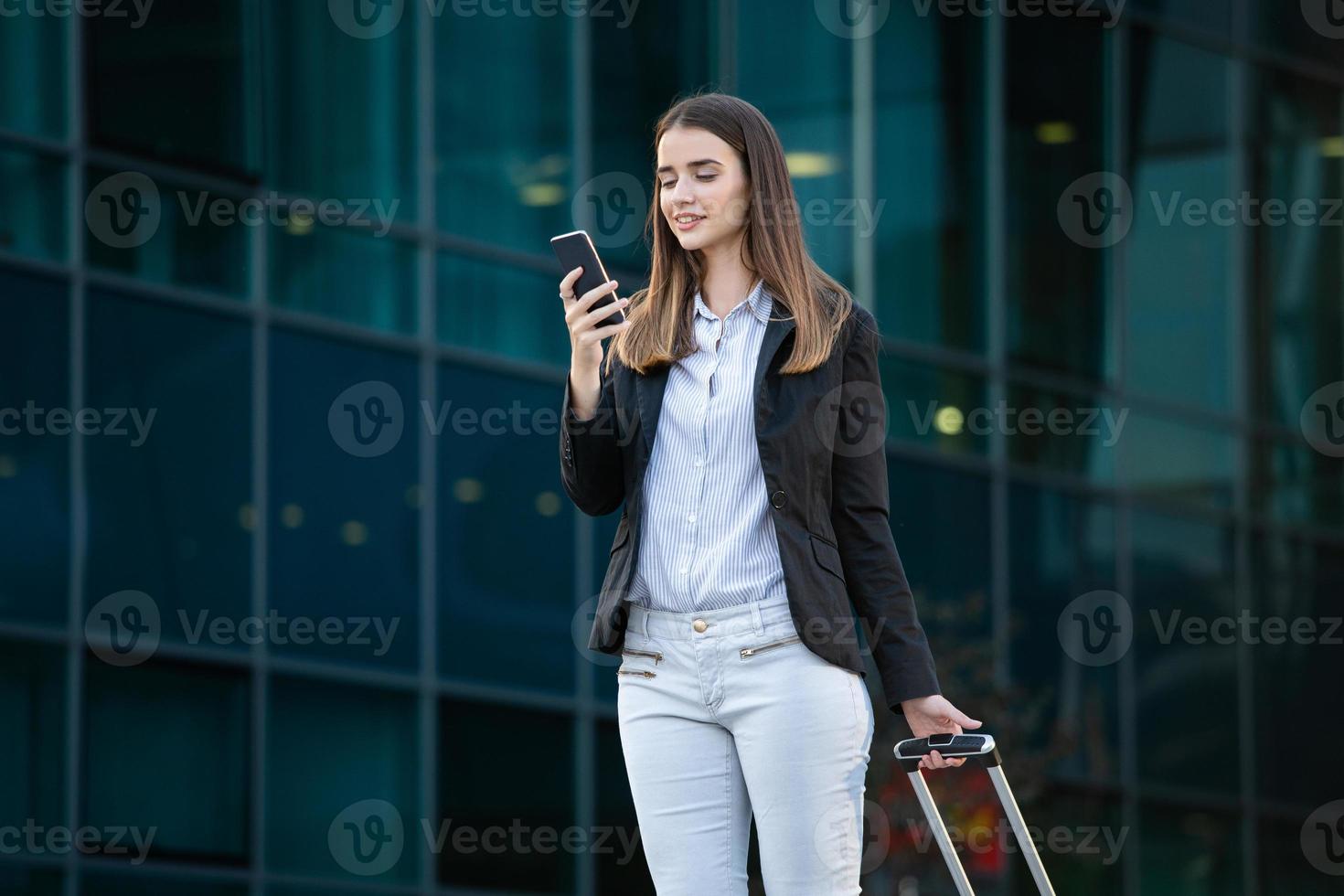 Pretty smiling female flight attendant carrying baggage going to airplane in the airport. Business woman at international airport looking at her mobile phone photo