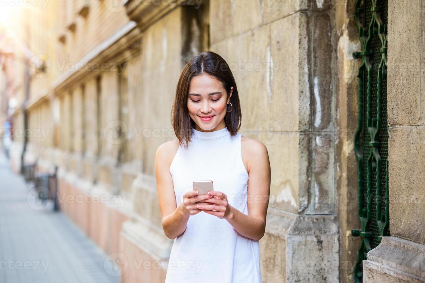 Beautiful asian woman using an application to send an sms message in her smartphone device. Happy young Asian woman with smartphone standing in the street photo