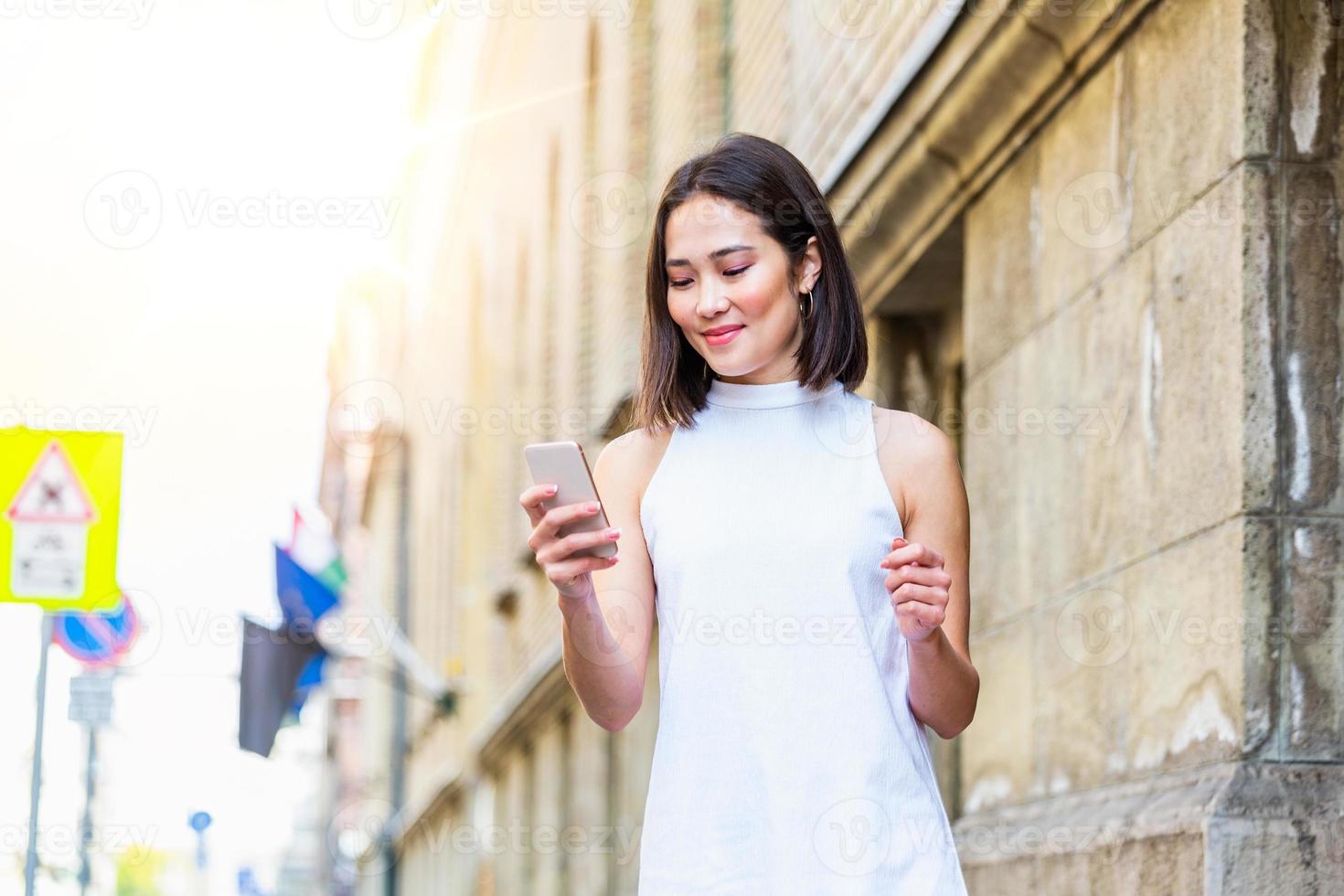 Beautiful asian woman using an application to send an sms message in her smartphone device. Happy young Asian woman with smartphone standing in the street photo