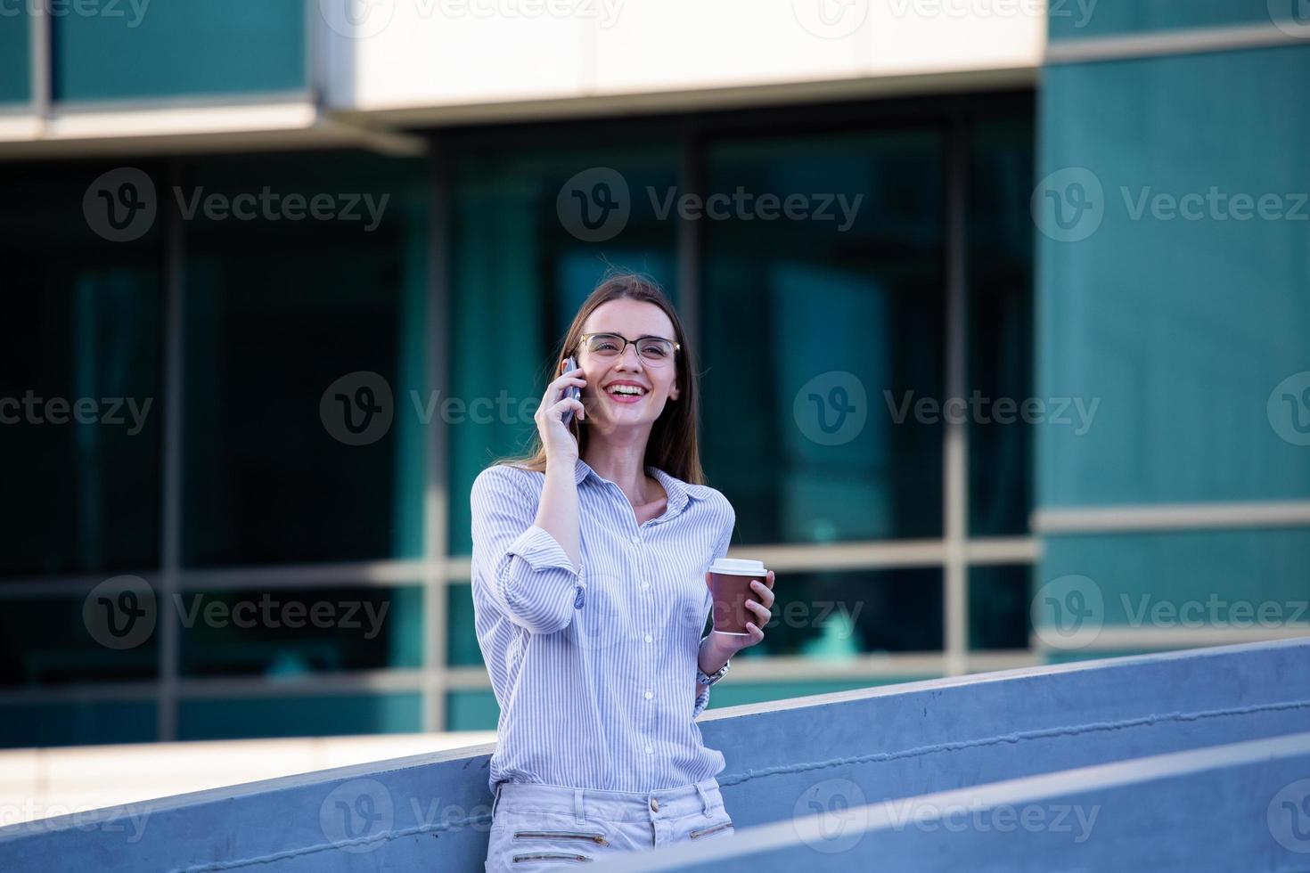 Executive business woman looking at mobile smartphone and drinking coffee from disposable paper cup in the street with office buildings in the background photo