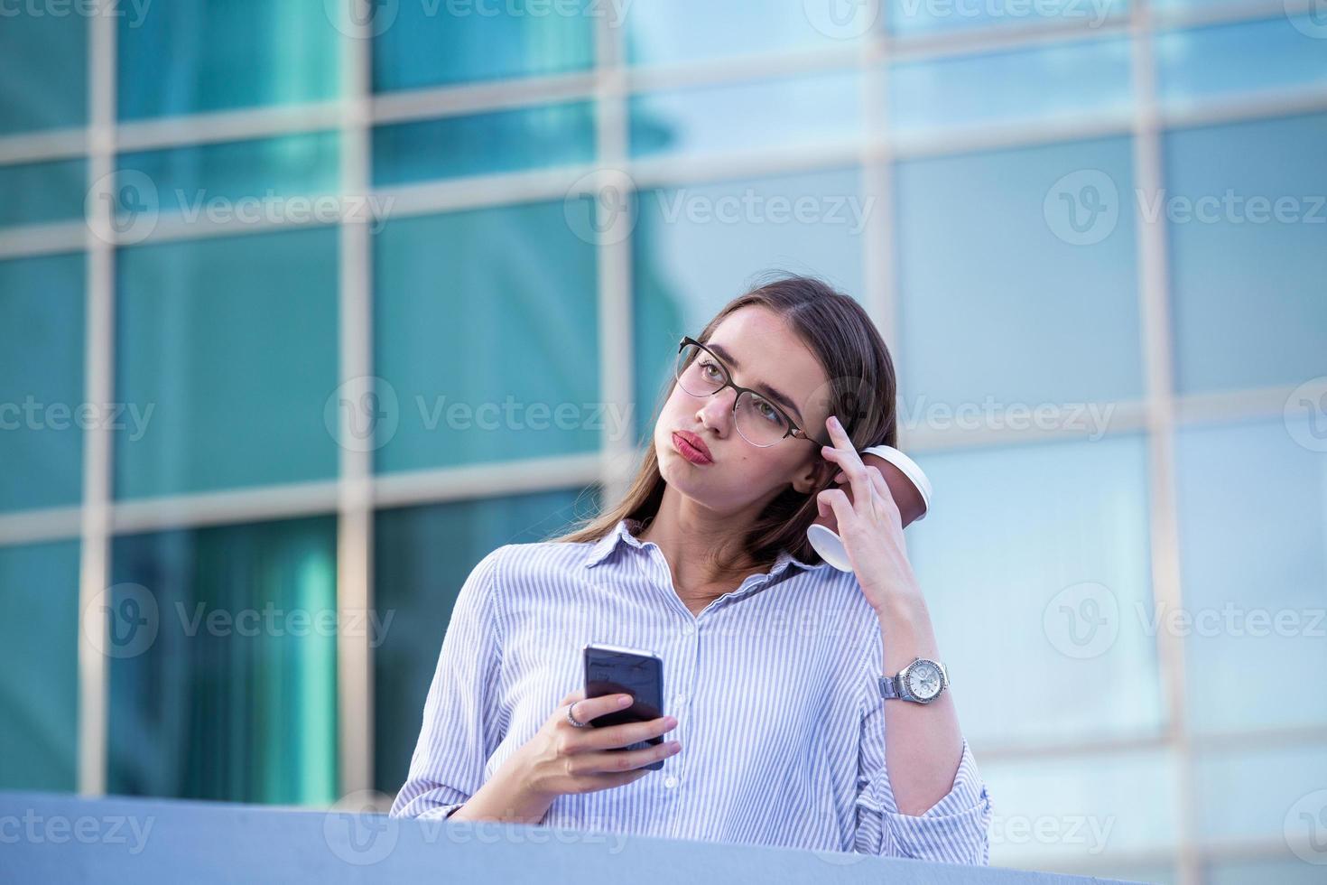 Business woman recieving bad news on smartphone and drinking coffee from disposable paper cup in the street with office buildings in the background photo