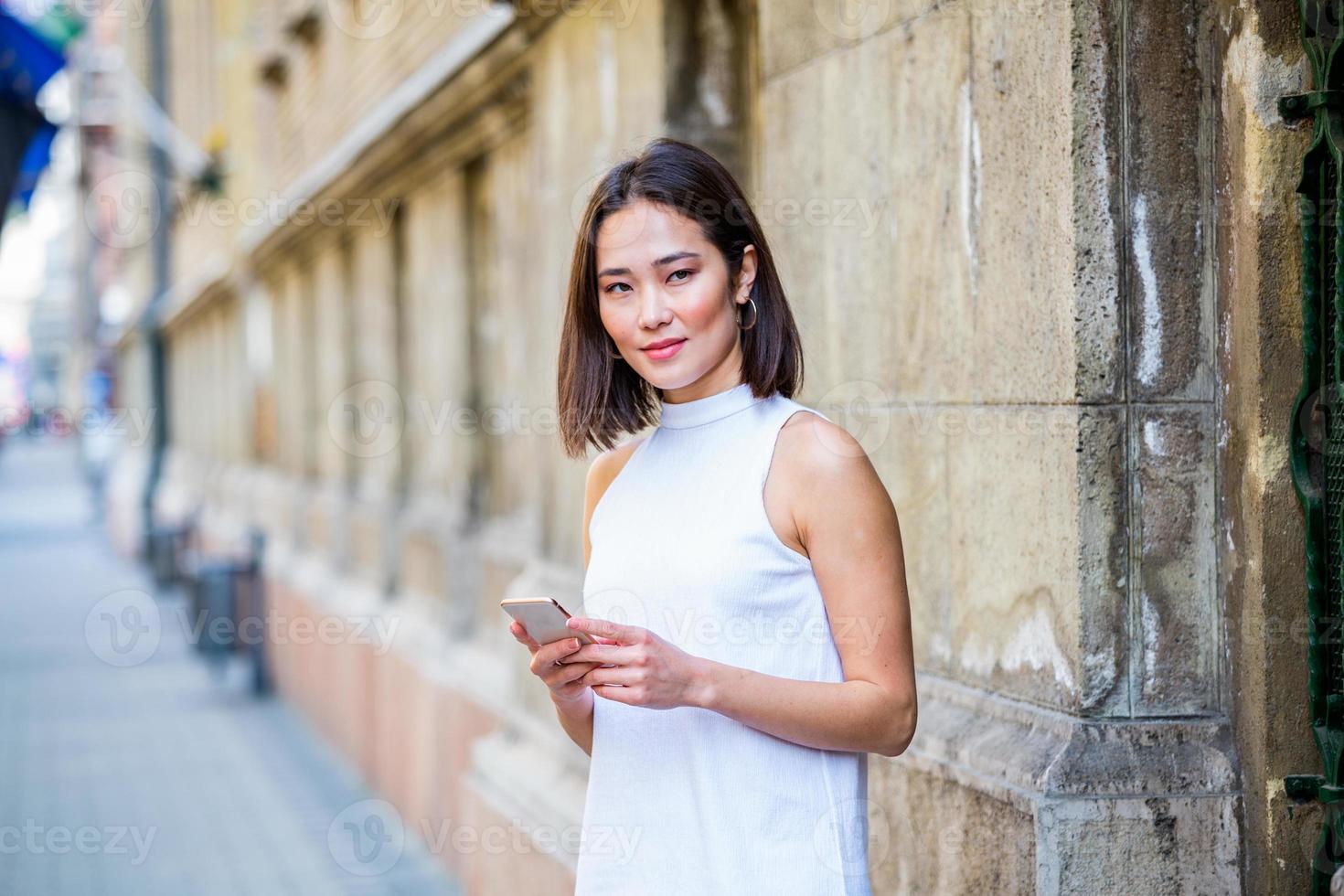 Beautiful asian woman using an application to send an sms message in her smartphone device. Happy young Asian woman with smartphone standing in the street photo