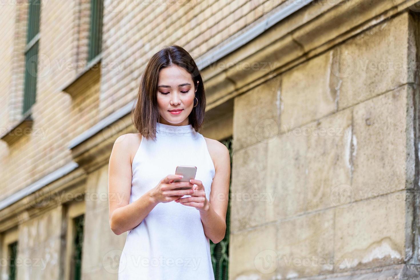 Beautiful asian woman using an application to send an sms message in her smartphone device. Happy young Asian woman with smartphone standing in the street photo