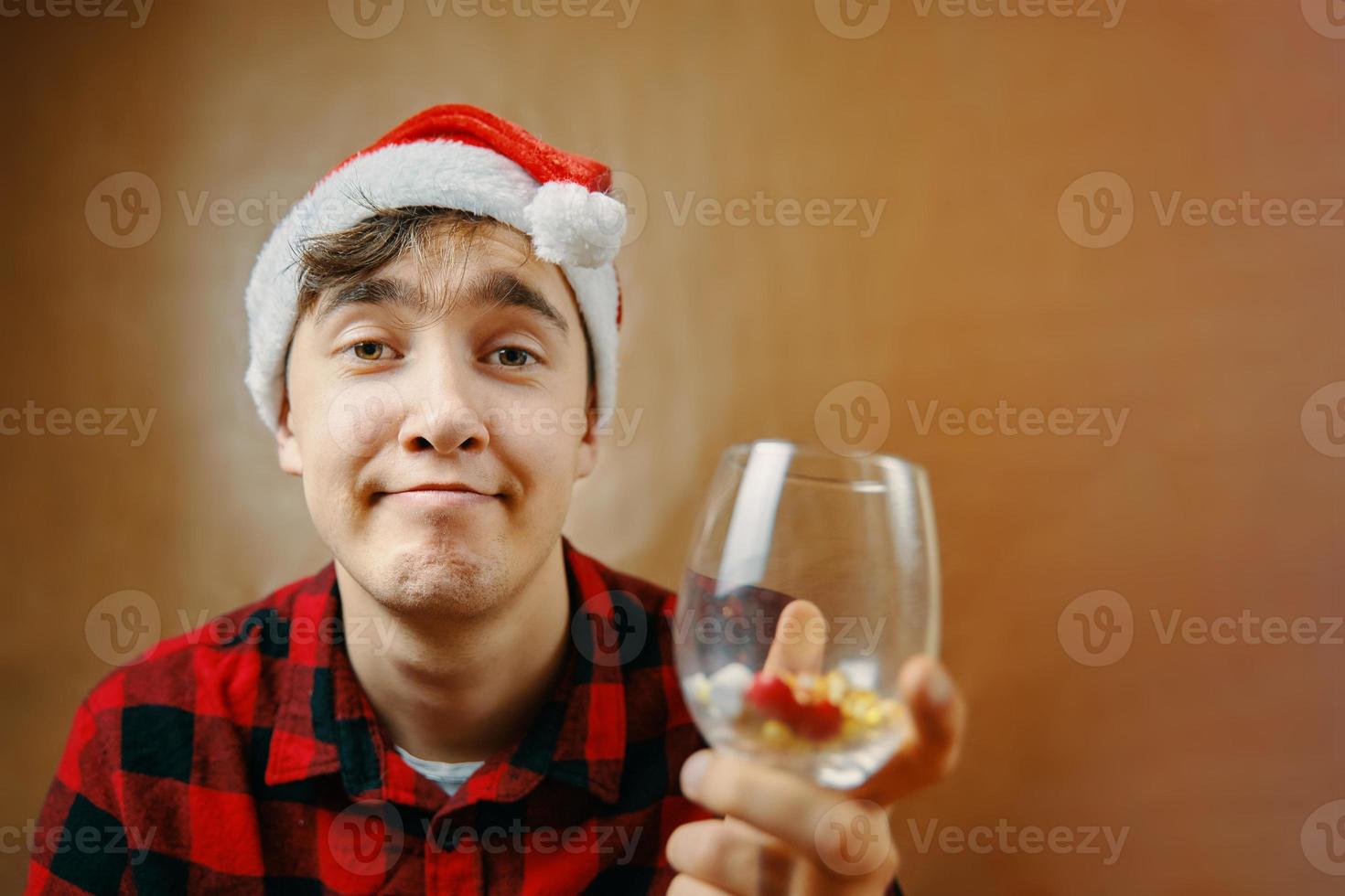 Guy in Santa Claus hat holds a glass with various pills. photo