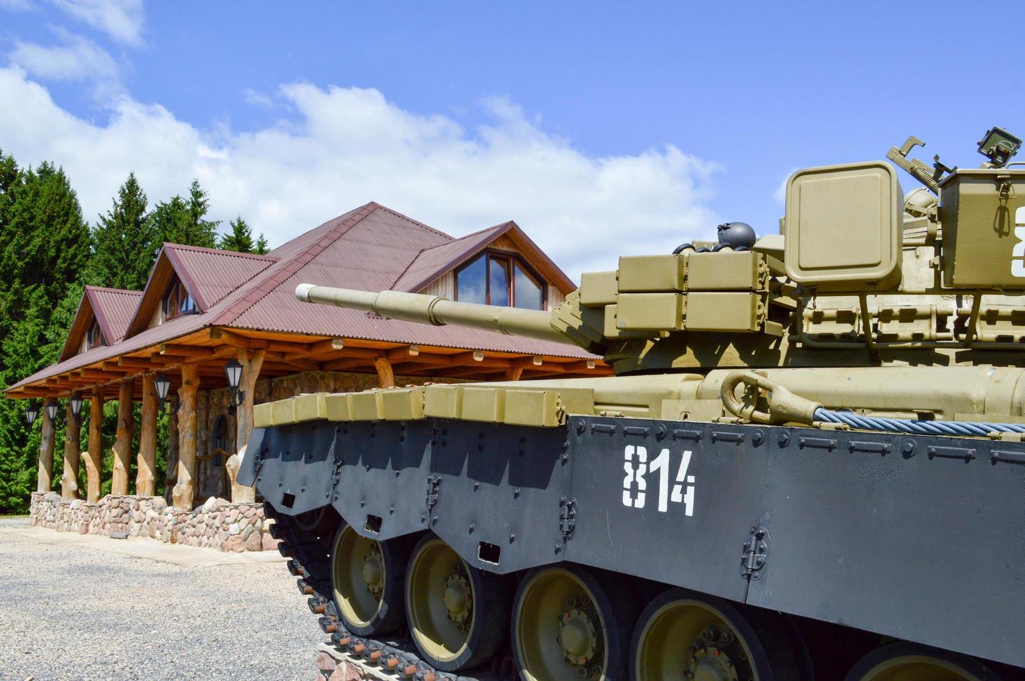 A large green military iron metal battle tank with a cannon is ridden parked next to the cottage house with a red tiled roof photo