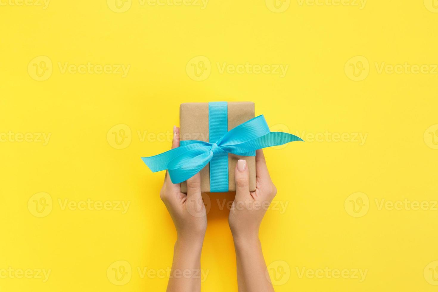 Woman hands give wrapped valentine or other holiday handmade present in paper with blue ribbon. Present box, decoration of gift on yellow table, top view with copy space photo