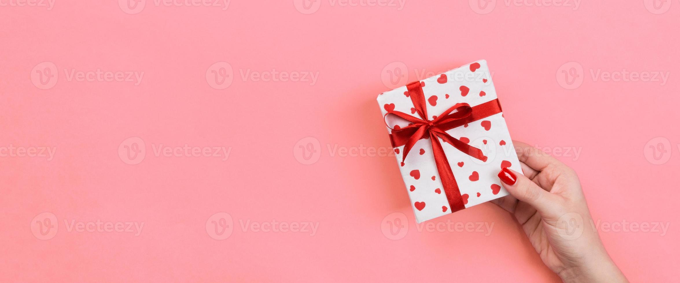 Woman hands give valentine or other holiday handmade present in paper with red ribbon. Present box, red heart decoration of gift on Coral color table, Top view banner with copy space for you design photo