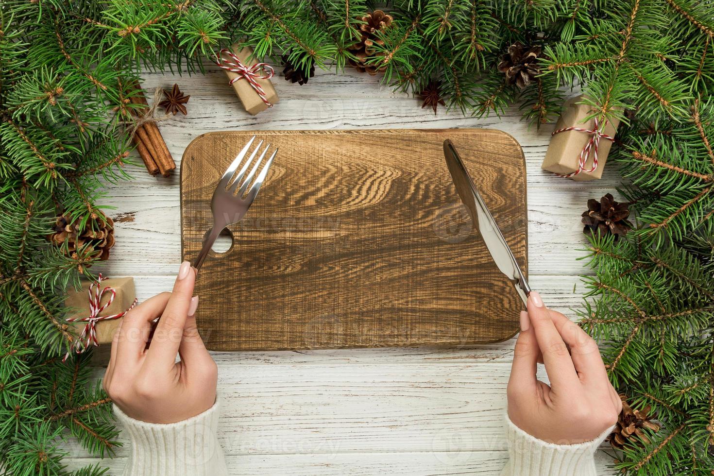 la chica de la vista superior sostiene un tenedor y un cuchillo en la mano y está lista para comer. placa rectangular de madera vacía sobre fondo de Navidad de madera. concepto de plato de cena navideña con decoración de año nuevo foto