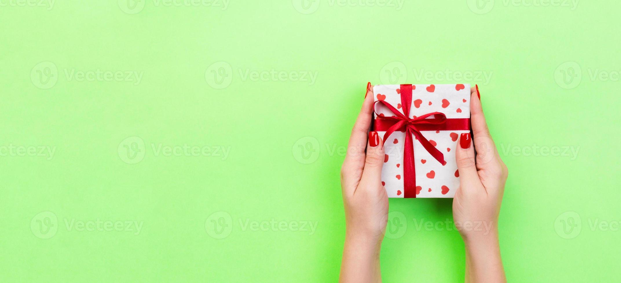 Woman hands give wrapped valentine or other holiday handmade present in paper with red ribbon. Present box, red heart decoration of gift on green table, Top view banner with copy space for you design photo