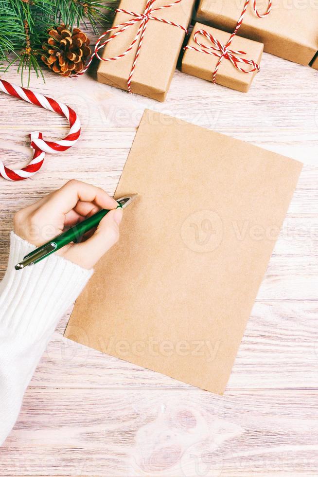 manos femeninas escribiendo una carta a santa claus. regalos de navidad con decoración navideña sobre un fondo de madera. vista desde arriba. tonificado foto