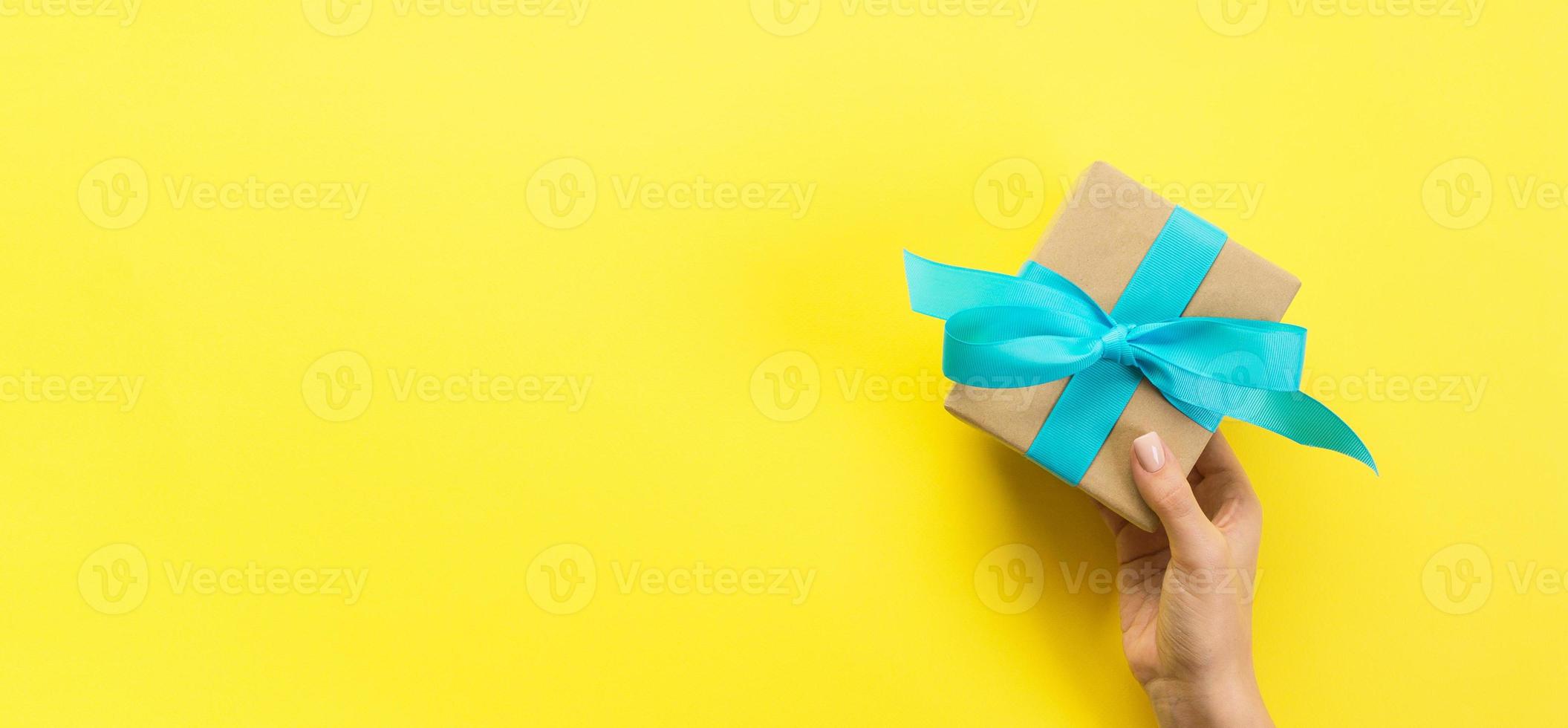 Woman hands give wrapped valentine or other holiday handmade present in paper with blue ribbon. Present box, decoration of gift on yellow table, Top view banner with copy space for you design photo