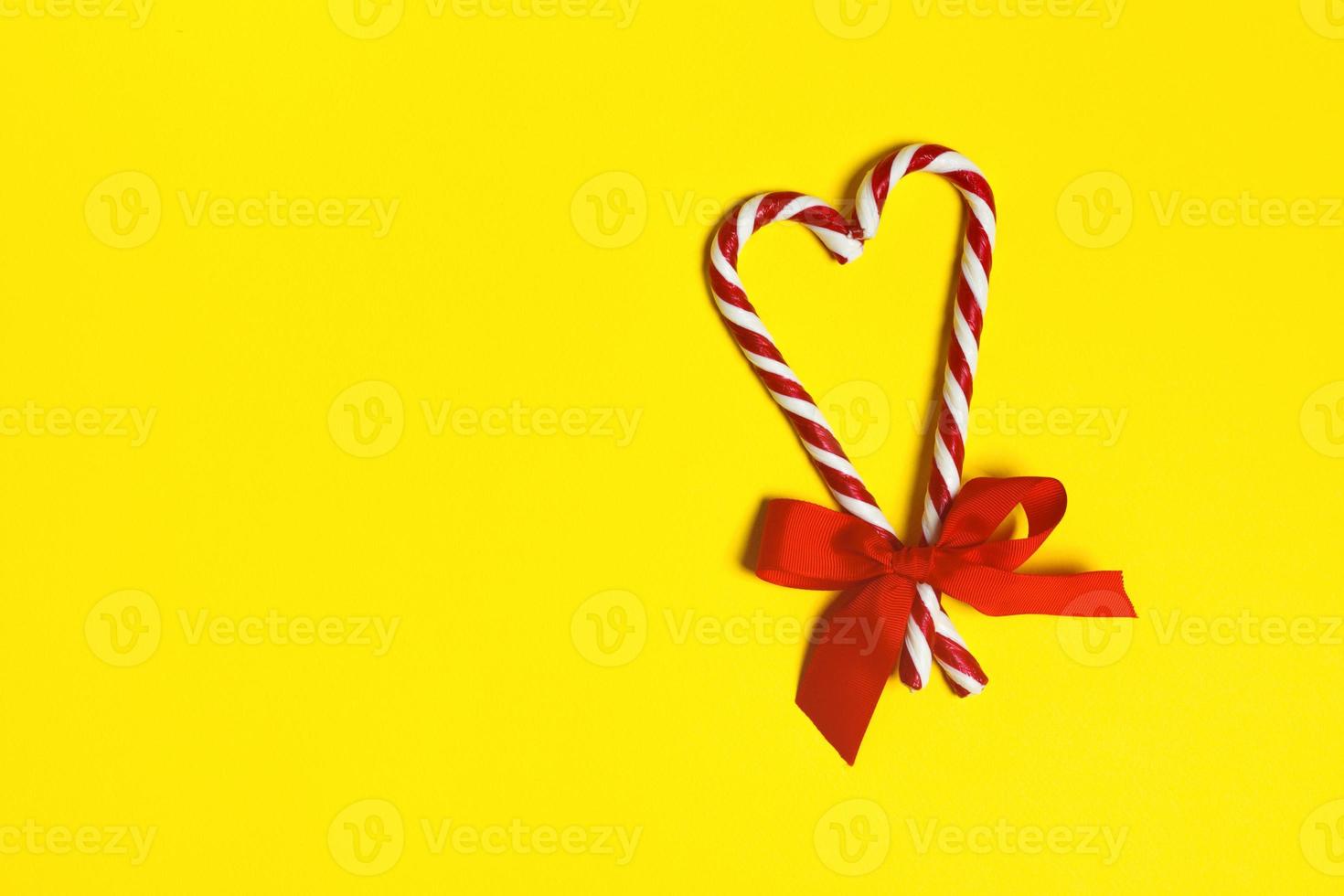 Two Christmas candy canes overlapping to form a heart and a red bow on a yellow background. copy space photo