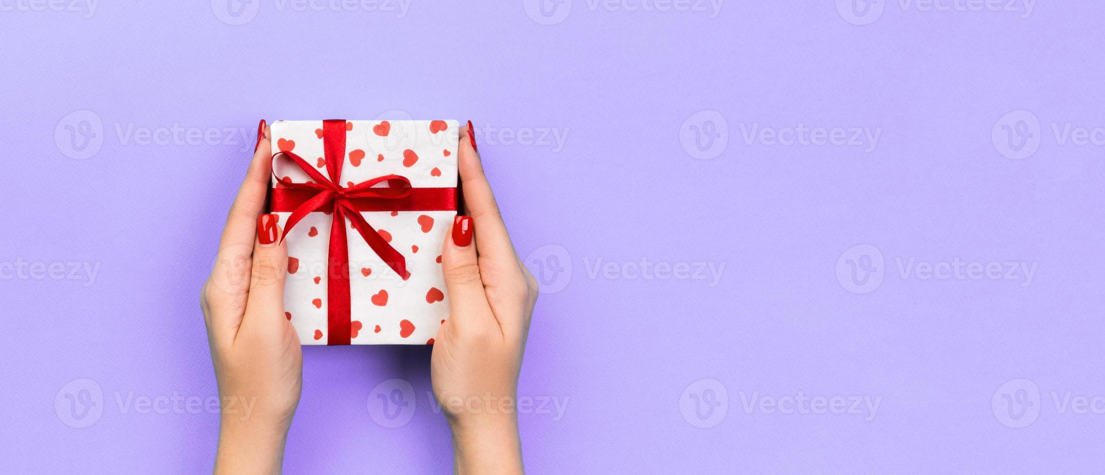 Woman hands give wrapped valentine or other holiday handmade present in paper with red ribbon. Present box, red heart decoration of gift on purple table, Top view banner with copy space for you design photo