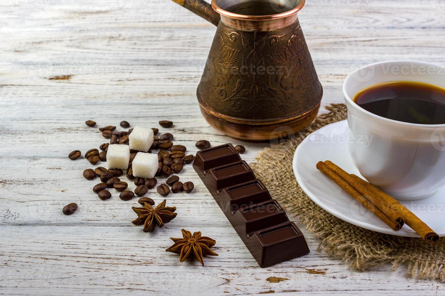 Coffee in a white cup on a small plate on sacking and cinnamon sticks. Coffee beans, pieces of sugar, anise and a chocolate bar on wooden table photo