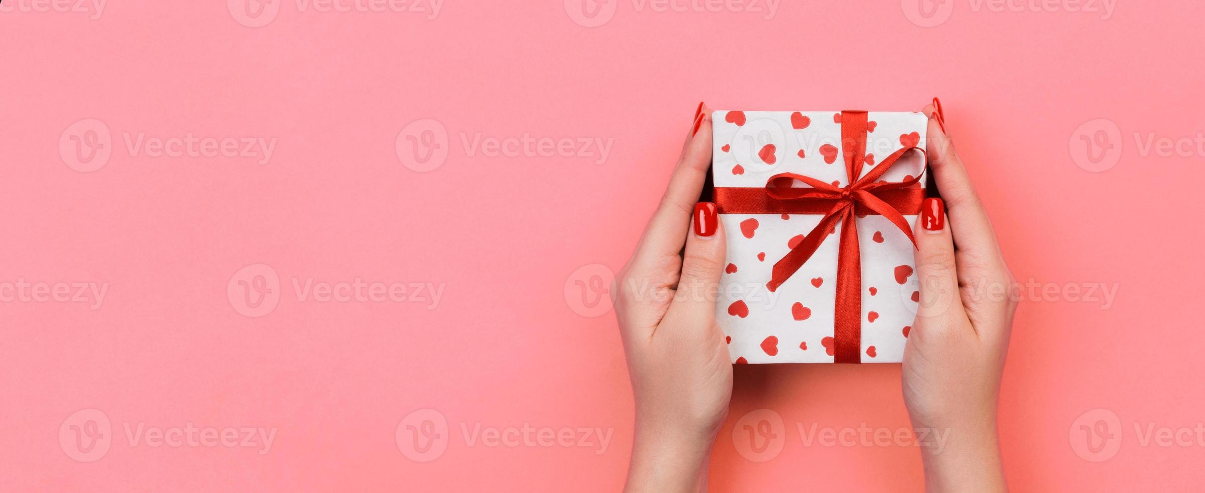 Woman hands give valentine or other holiday handmade present in paper with red ribbon. Present box, red heart decoration of gift on Coral color table, Top view banner with copy space for you design photo