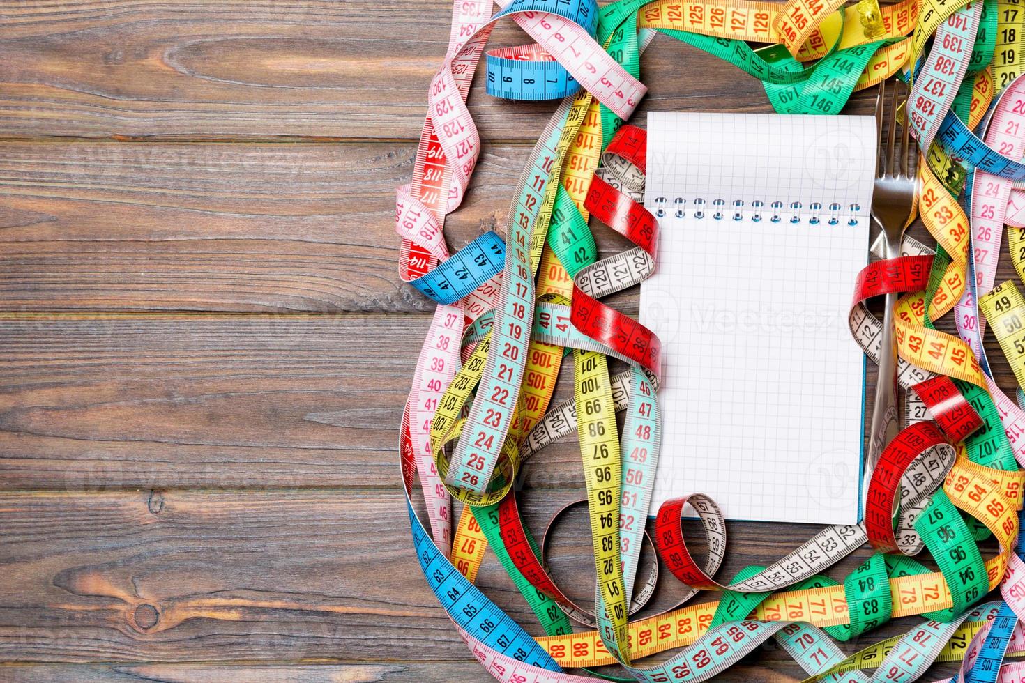 Fitness concept with empty space for your design on dark wooden background. Top view of notepad and fork surrounded with colorful measure tapes photo