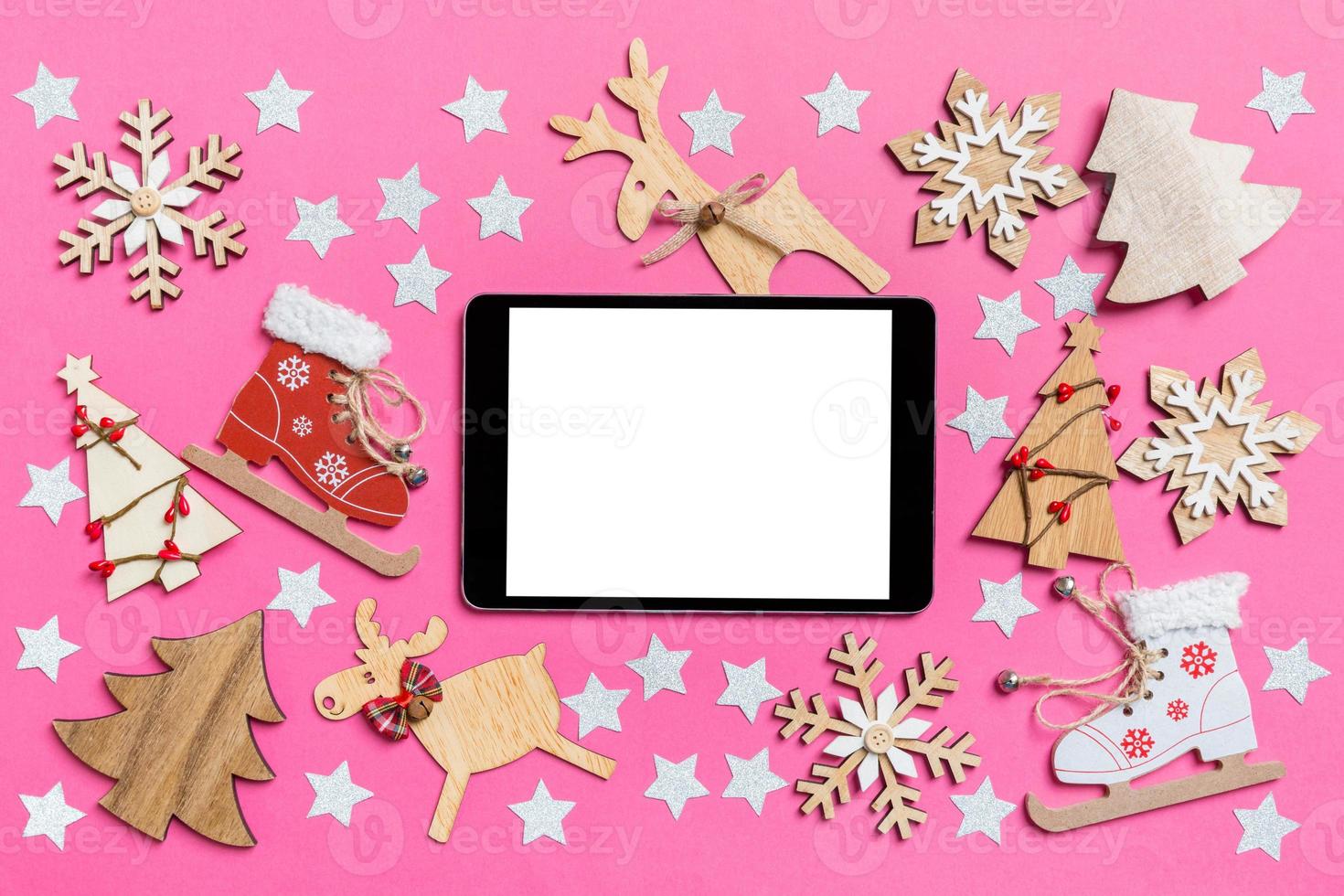 Top view of digital tablet, pink background decorated with festive toys and Christmas symbols reindeers and New Year trees. Holiday concept photo