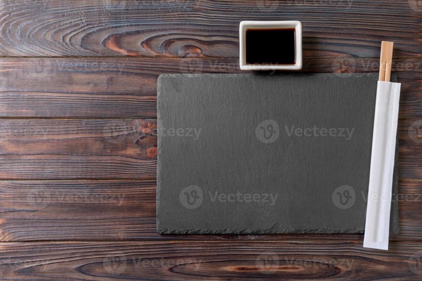 Empty rectangular black slate plate with chopsticks for sushi and soy sauce on wooden background. Top view with copy space photo