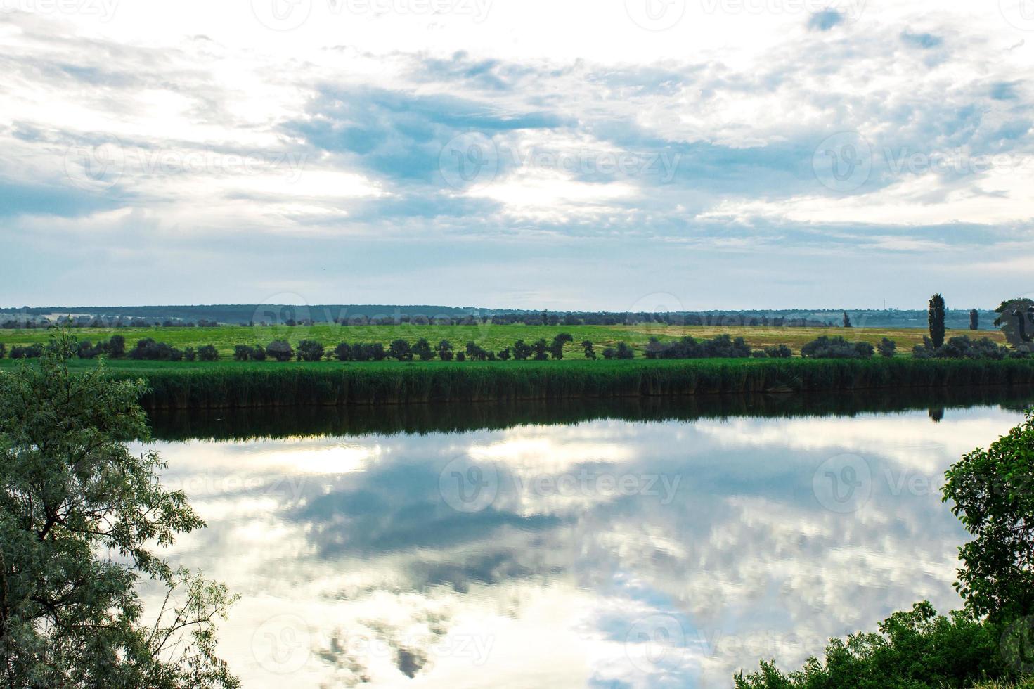 Summer evening dawn sky clouds reflected on a lake of water on the coast with green grass and forest trees photo