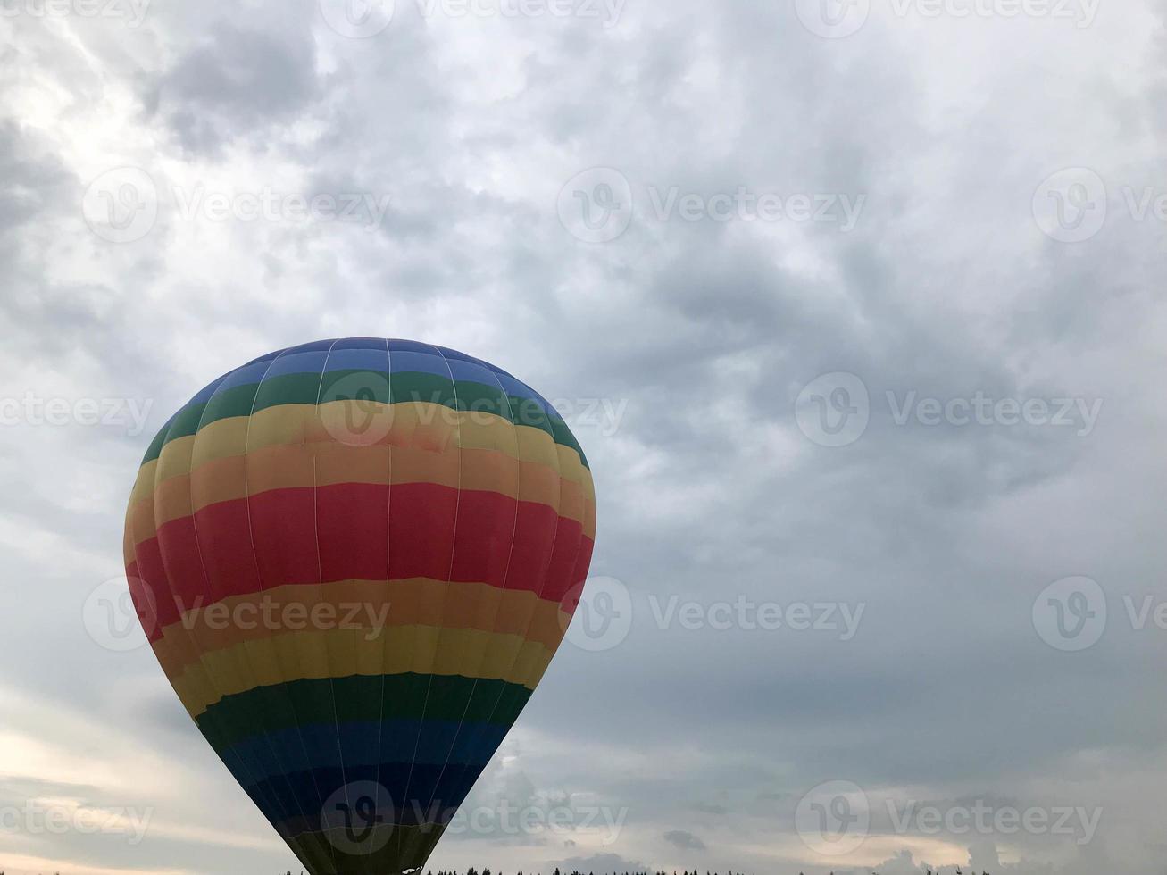 Large multi-colored bright round rainbow colored striped striped flying balloon with a basket against the sky in the evening photo