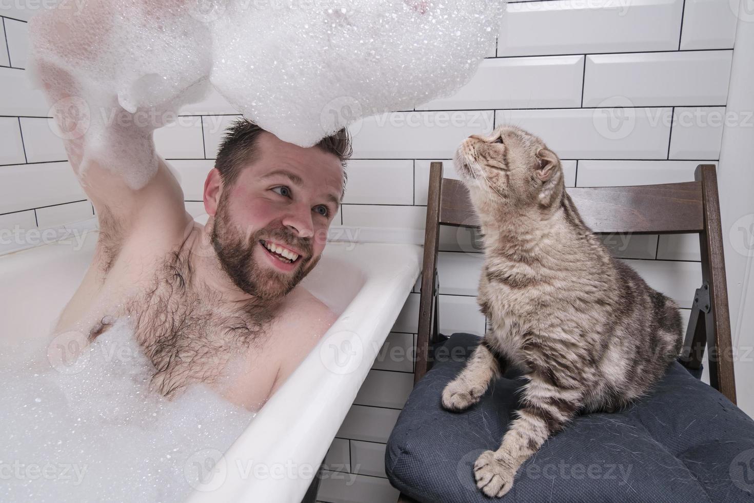 Satisfied man bathes in a bathtub, he holds on hand a lush soap suds, which his adored cat is interested in. photo