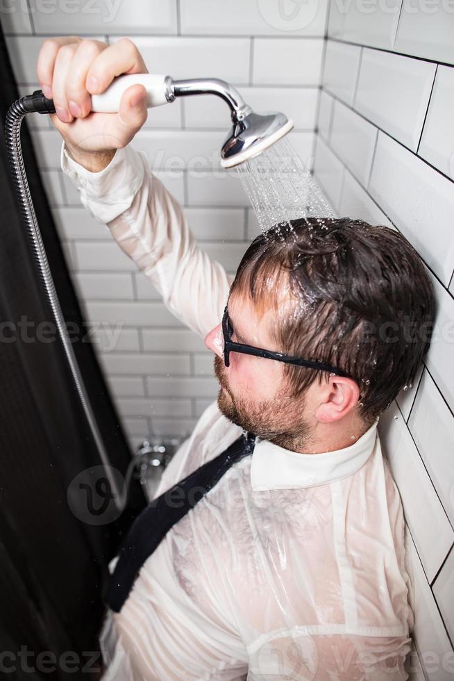 Man in stress after work. He stands in the bathroom in glasses and clothes, pouring running water over himself from a watering can. photo