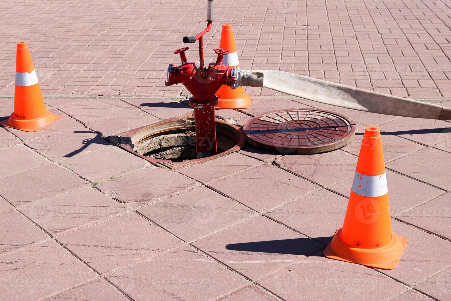 A large red metal fire hydrant with hoses and sleeves connected to the water source in the sewer hatch and emergency triangular orange cones warning of the danger photo
