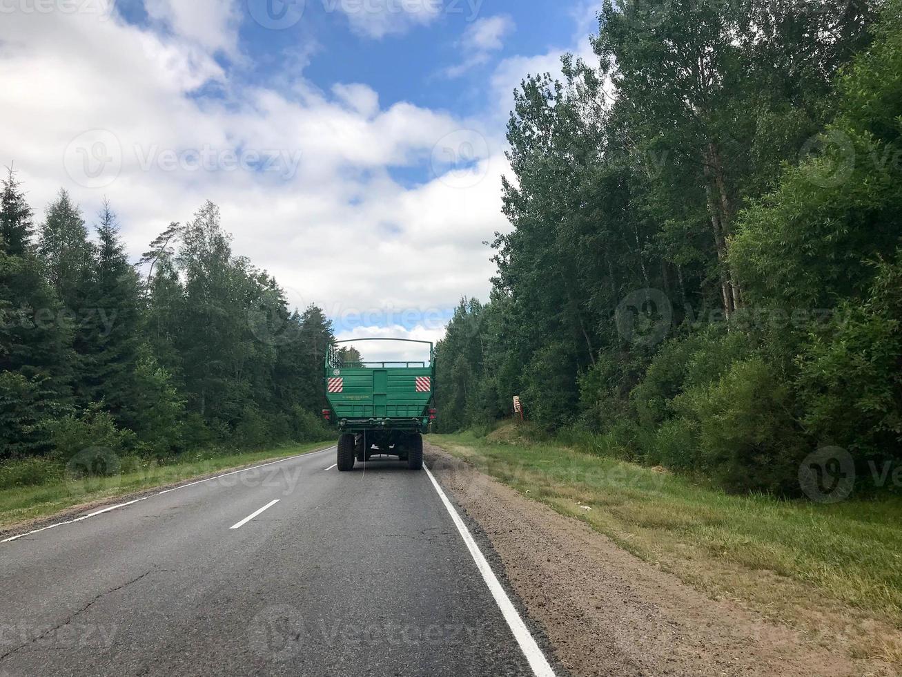 A truck, a tractor with a large green trailer is driving along a forest asphalt road with green trees on the grounds photo