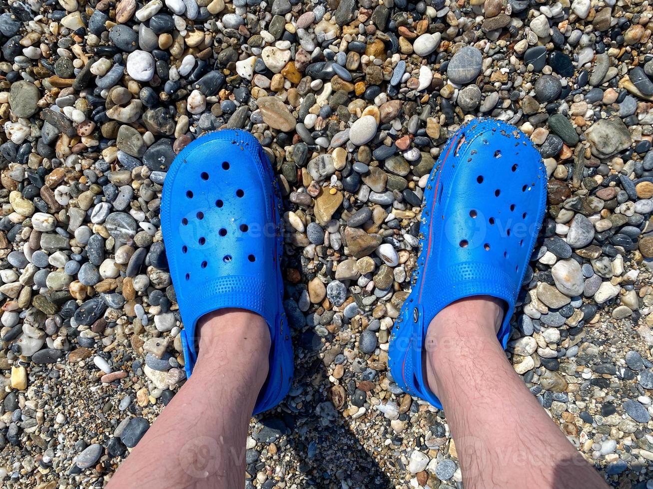 piernas de hombres, pies en zapatillas de goma azul contra el fondo de guijarros y arena en unas vacaciones en la playa en un cálido paraíso tropical del este del país resort sureño foto