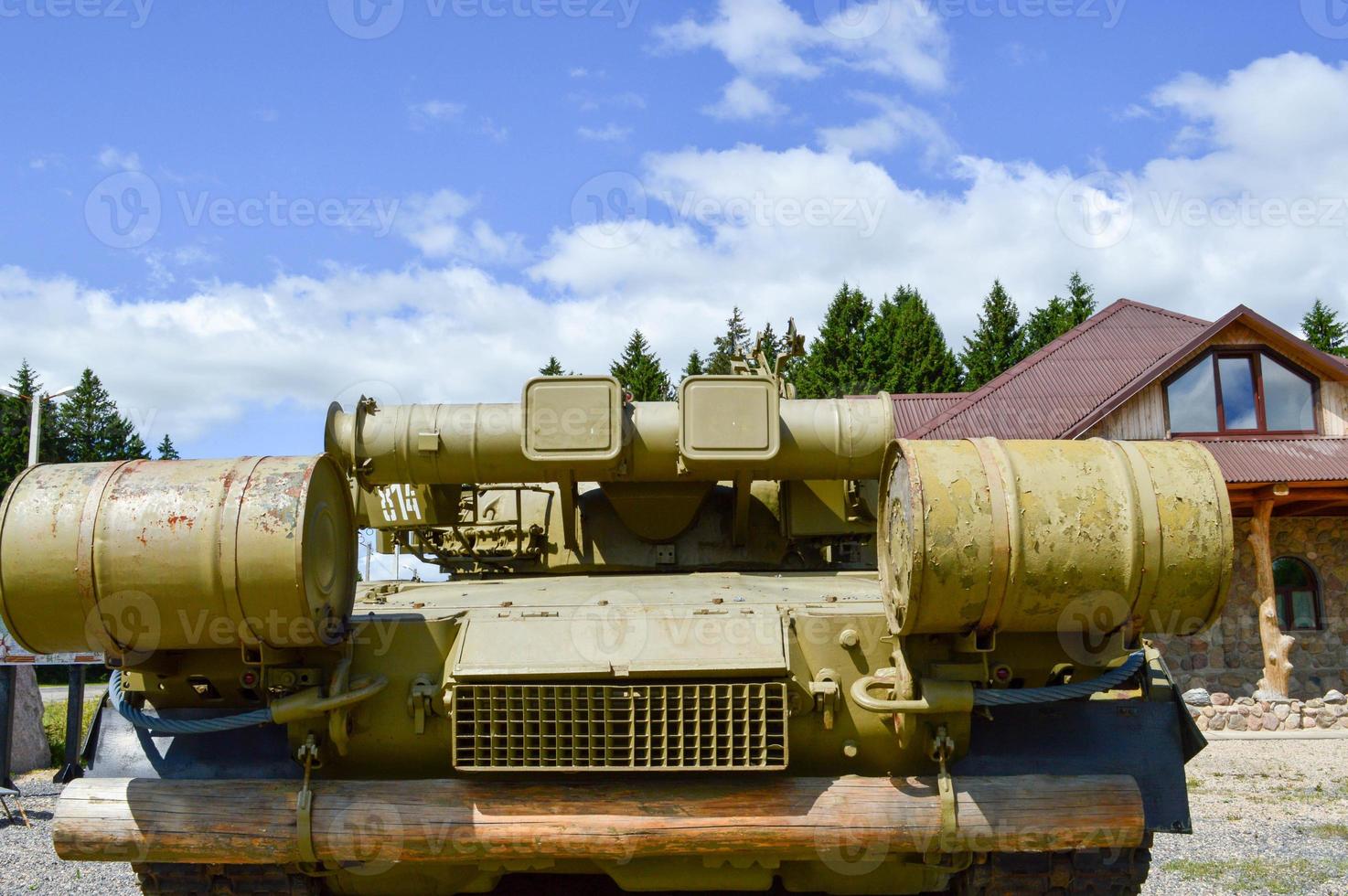 A large green military iron metal battle tank with a cannon is ridden parked next to the cottage house with a red tiled roof photo