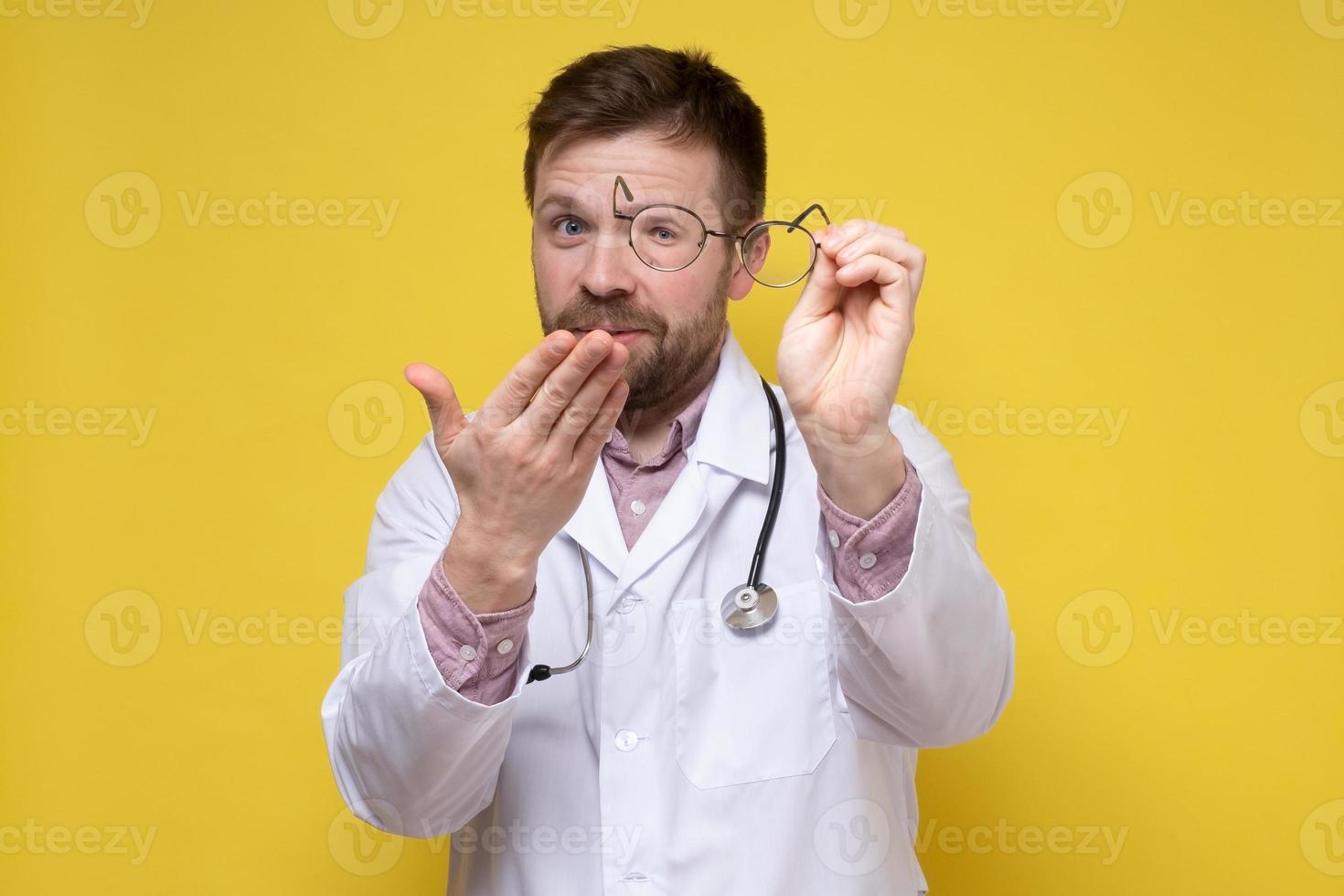 Doctor in a white coat smiles in surprise, makes a gesture with hand and looks with one eye through the lens of glasses. photo