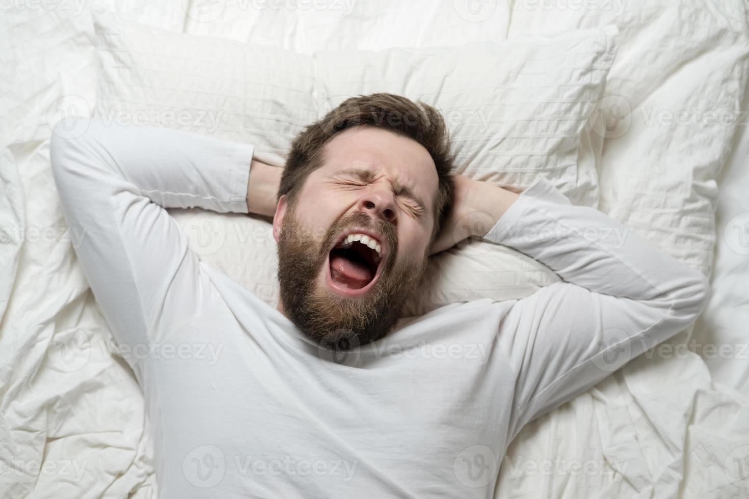 Cute bearded man wakes up from sleep, he yawns and stretches as he lies in bed. Top view. Close-up. photo