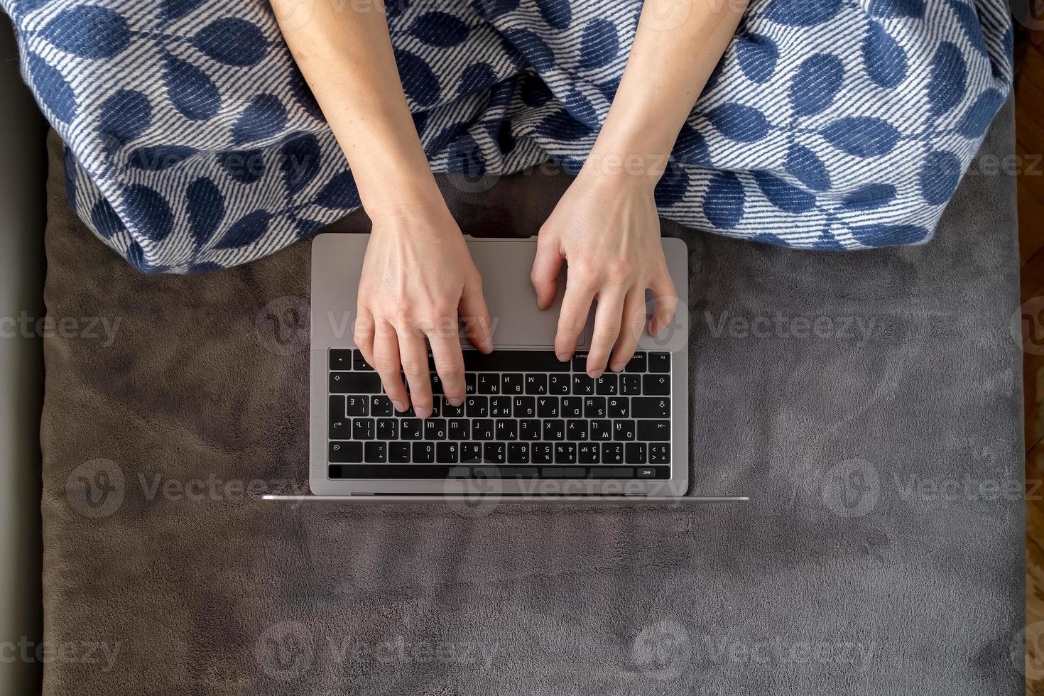A woman wrapped in a plaid works with a laptop sitting on a soft sofa at home. photo