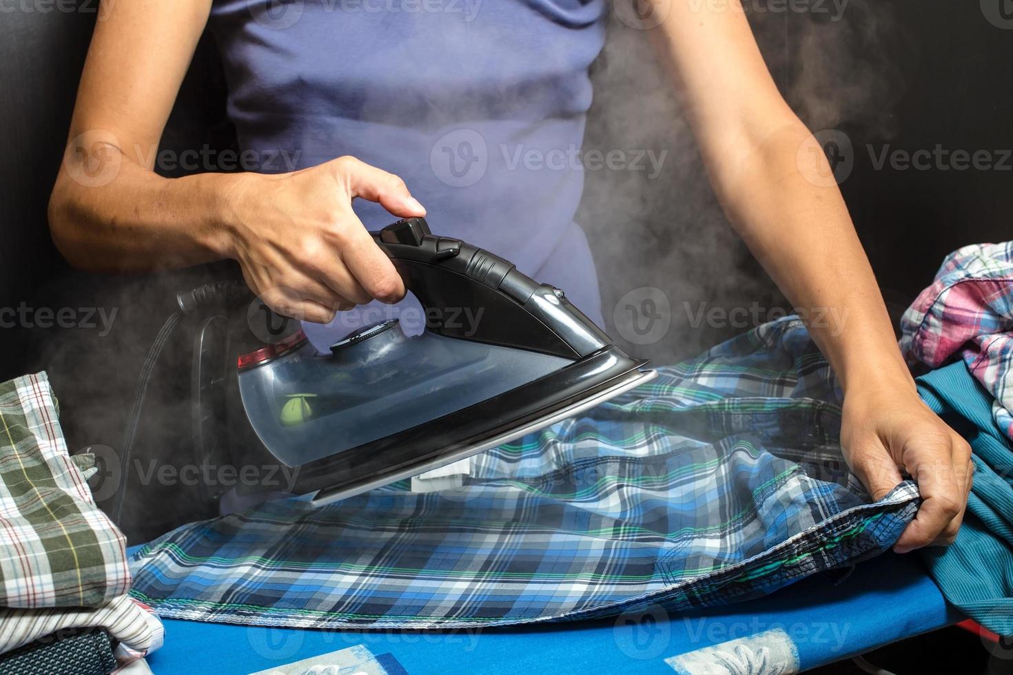 Woman is ironing clean clothes with a hot iron and steam, neatly folded shirts lie nearby. Household chores. photo