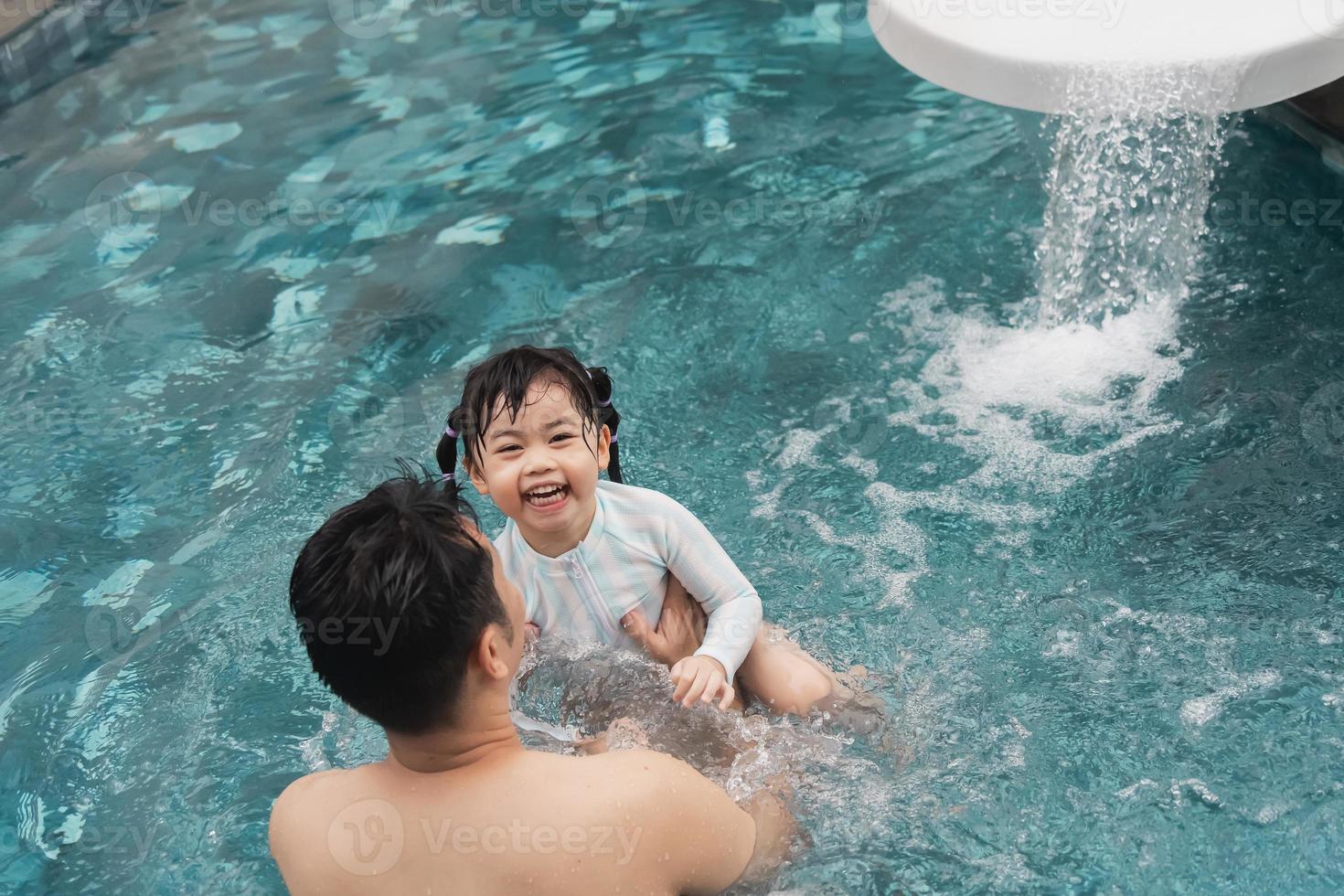 padre e hija asiáticos nadando jugando al tobogán en la piscina del resort, sonriendo y riendo. divirtiéndose en la piscina del hotel resort, concepto familiar feliz. foto