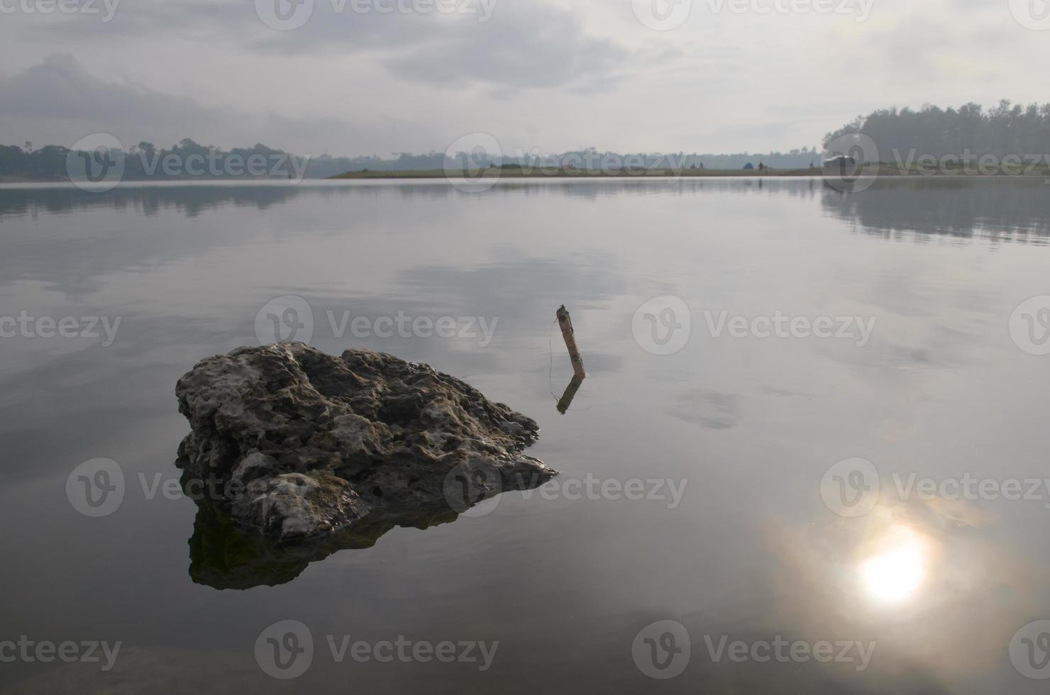 Front view of a large rock in Karangkates lake Indonesia with calm water conditions at sunrise with a mountains background and a foggy sky photo