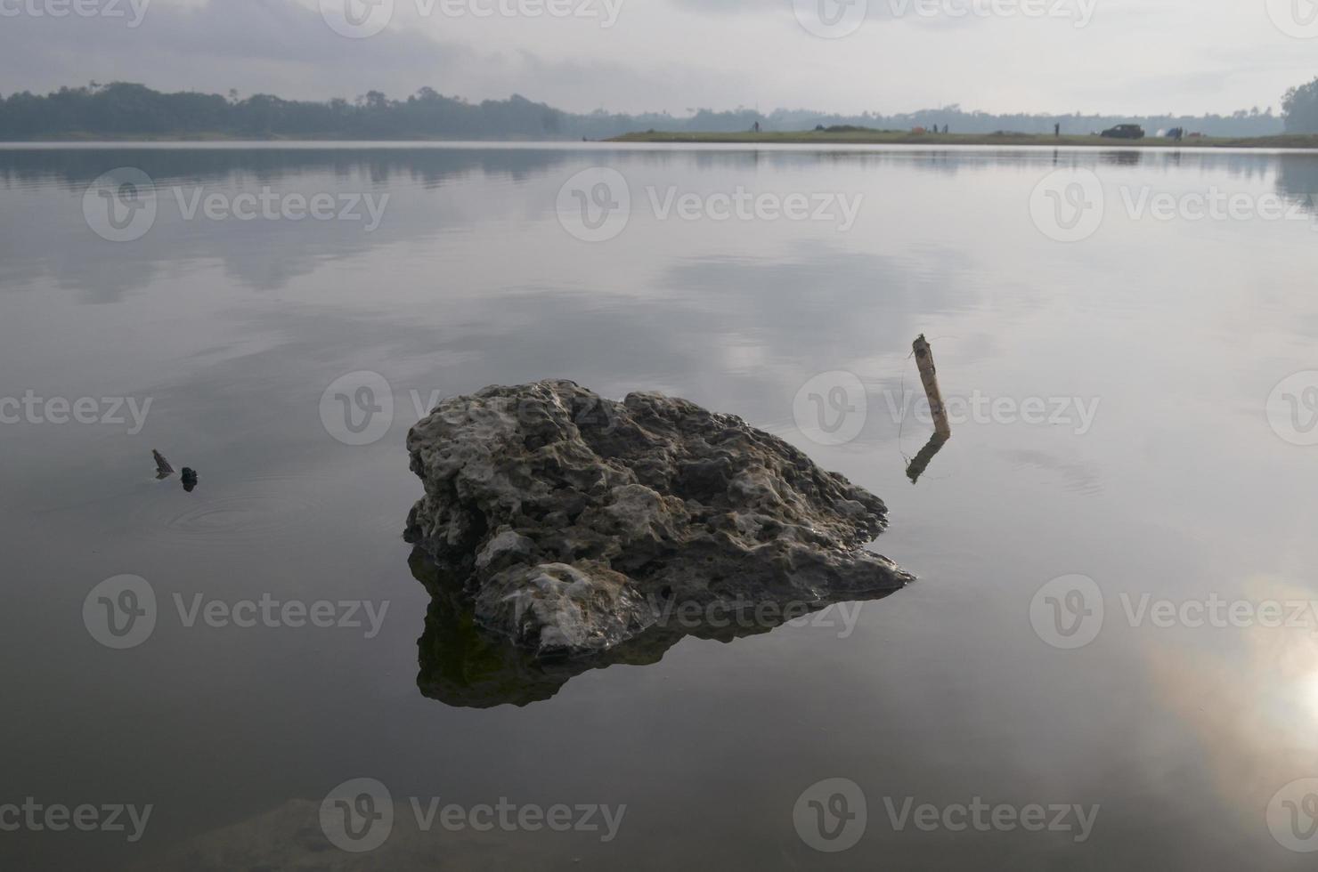 Front view of a large rock in Karangkates lake Indonesia with calm water conditions at sunrise with a mountains background and a foggy sky photo