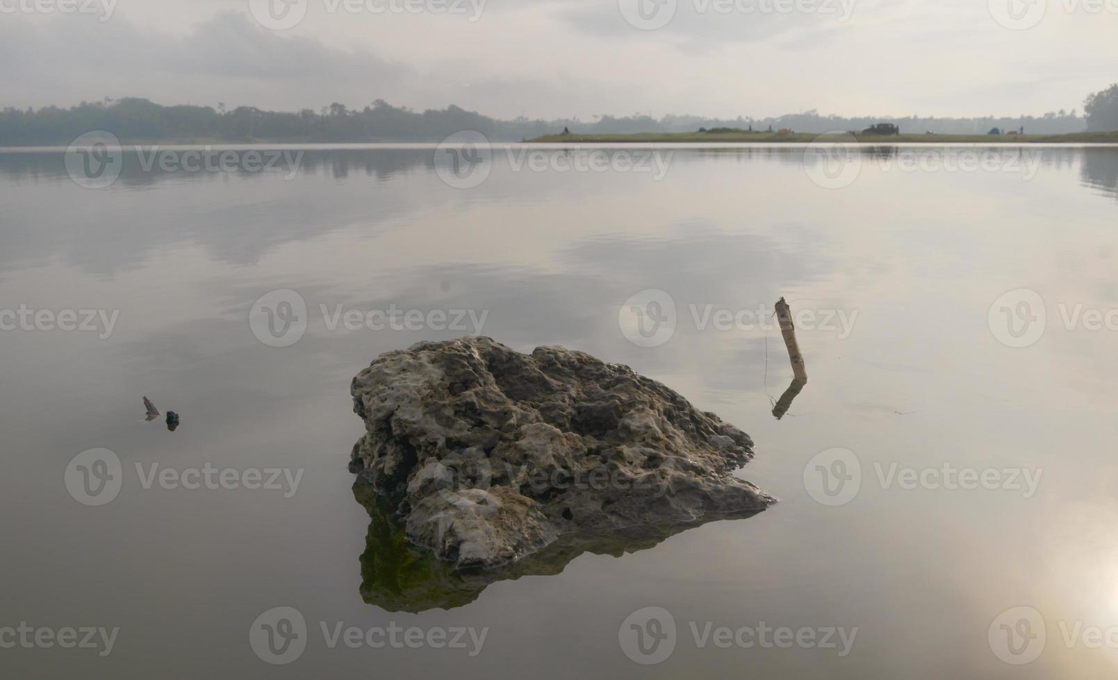 Front view of a large rock in Karangkates lake Indonesia with calm water conditions at sunrise with a mountains background and a foggy sky photo